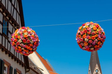 Parlak renkli çiçek aranjmanları kaldırım taşı sokağının yukarısındaki iplerden sarkıyor ve pitoresk köyde şenlikli bir atmosfer yaratıyor. Açık mavi gökyüzü neşeli bir zemin sağlar.