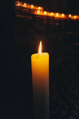 Tek bir mum parlak bir şekilde yanar, loş bir alanda sıcak bir parıltı bırakır. Arka planda, birkaç mum daha yavaşça titreşiyor, sükunet ve yansıma ortamı yaratıyor..