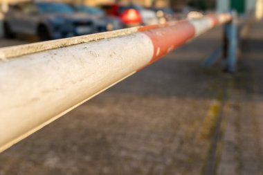 Kentsel bir ortamın girişinde kırmızı ve beyaz bir bariyer duruyor, arka planda birkaç park etmiş araç temiz güneş ışığı altında. Sahne günlük aktivitenin bir anını yakalıyor..