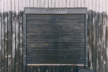 Yıpranmış metal bir kepenk ahşap bir pencere çerçevesini kaplıyor. Yaş ve ihmal belirtileri gösteriyor. Ayar, öğleden sonra saatlerinde çekilen kentsel bir ortamı yansıtıyor..