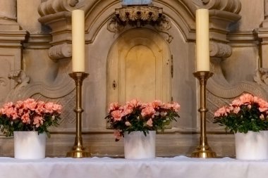 Huzurlu bir kilise sunağında uzun mumların yanında zarif pembe çiçek aranjmanları bulunur. Yumuşak ışıklandırma ibadet sırasında huzurlu atmosferi arttırır..