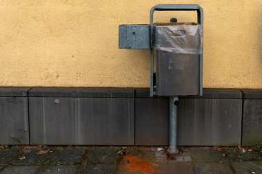 Bej bir duvara dayalı metal bir çöp konteynırı odak noktasını yakalıyor. Şeffaf bir plastik kaplama içeriyor ve aşınma izleri gösteren bir kaldırımda duruyor..