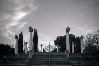Adımlar, akşam ışığı altında bulutlu bir gökyüzünün altında yer alan uzun ağaçların arasında yer alan küresel sütunlarla süslenmiş çarpıcı bir yapıya doğru uzanır..