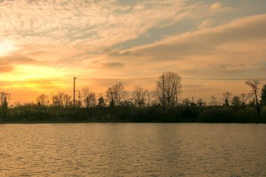 Yumuşak ışık, arka planda güneş batarken sakin göl üzerinde altın renkler bırakır. Ağaçların siluetleri alacakaranlık sırasında suyun kenarında huzurlu bir atmosfer yaratır..