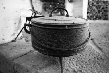 Eski bir dökme demir tencere kırsal bir mutfaktaki yıpranmış taş bir yüzeye dayanır. Esrar, konforlu bir alanda geleneksel pişirme araçlarıyla çevrelenmiş, yaş ve kullanım belirtileri gösteriyor..