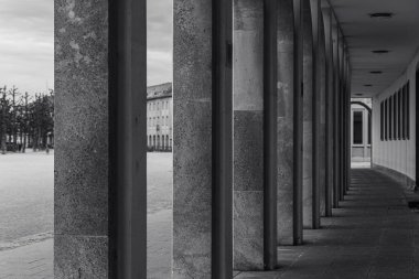 Eski bir binanın uzun, gölgeli koridorunda kaldırıma gölge düşüren taş sütunlar var. Sahne, tarih ve sessiz bir yansımayı çağrıştırıyor..