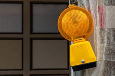 Parlak sarı bir uyarı ışığı bir inşaat alanının yanında asılı duruyor ve yakındaki şehir binasına parlıyor. Olay yeri, açık gökyüzünün altındaki yoğun bir alanda devam eden bir çalışmayı gösteriyor..