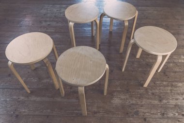 Beş basit ahşap tabure, yaşlanmış ahşap bir zeminde dairesel bir düzende konumlandırılmıştır. Minimalist bir tasarım sergilenmektedir..