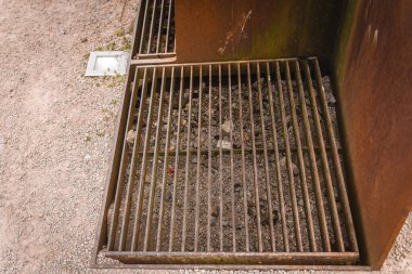 Paslı bir metal ızgara kayalar ve toprak da dahil olmak üzere bir enkaz yığınının üzerinde, açık hava ortamındaki çakıl yolunun yanında bulunur. Ayarlar yıpranmış ve ihmal edilmiş görünüyor..