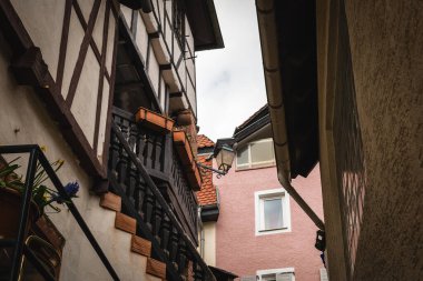 Dar bir sokakta gezinen ziyaretçiler, çiçek kutularıyla süslenmiş yarım ahşap evlere hayran kalıyorlar. Gökyüzü bulutlu, köyün sıcak atmosferine katkı yapıyor..