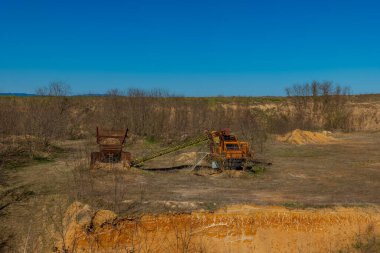 Paslı maden makineleri, temiz mavi gökyüzünün altındaki ıssız ve kuru bir alanda bozulmamış bir şekilde duruyor. Çevresindeki manzara, kahverengi toprak ve seyrek ağaçlarla kaplıdır..