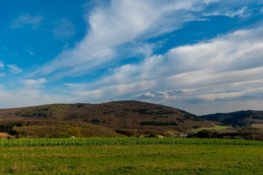 Yuvarlanan yeşil tepeler, parlak mavi bir gökyüzünün altında, öğleden sonra ışığında süzülen ince bulutlarla birlikte arazi boyunca uzanıyor. Kırsalda barışçıl bir atmosfer hüküm sürüyor.