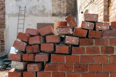 Yığılmış tuğlalar kısmen inşa edilmiş bir duvarın önünde, üzerine yaslanmış bir merdiven var. Güneş ışığı, gün içinde tuğlaların dokularını ve renklerini vurgular.