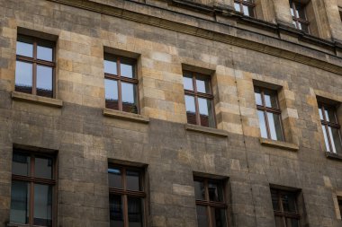 Kahverengi ve bej renkli taşlar eski bir binanın dış yüzeyini oluşturur. Simetrik pencereleri güneş ışığını yansıtır. Yapı, şehirdeki tarihi mimari güzelliğin bir kanıtı olarak duruyor..