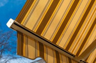 Güneşli bir alan boyunca uzanan güneşli bir tentenin altın çizgileri gölgeler yerde dans ederken ve ağaçlar hafif esintide sallanırken rahat bir ortam yaratıyor..