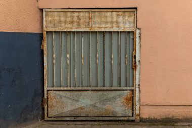 Dikey parmaklıkları olan yıpranmış metal bir kapı desenli bir duvara dayanır. Pas yaş belirtilerini gösteriyor, şehir manzarasına karakter katıyor..