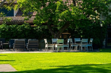 Sandalyeler ve masalar, yeni biçilmiş çimenlere davetkar bir şekilde oturur ve doğa tarafından çevrelenmiş sakin bir açık hava ortamında arkadaşlara huzurlu bir buluşma yeri sunar..
