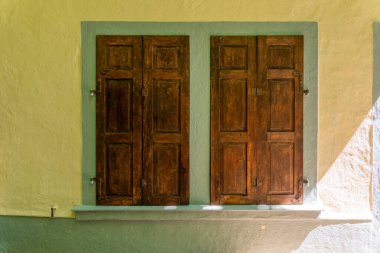 Güneş ışığı, büyüleyici sarı bir duvarda dans eder. Özenle hazırlanmış ahşap panjurlar içerir. Sahne sükunet ve kırsal zarafeti, dinginliği ve sıcaklığı simgeliyor..