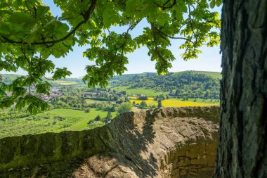 Taze yeşil yapraklardan oluşan bir gölgeliğin altında, kayan tepelerin panoramik manzarası açılır, çiçek açan tarlalar ve tuhaf köyler sergilenir, sakin bir bahar gününde parlak güneş ışığıyla aydınlatılır..