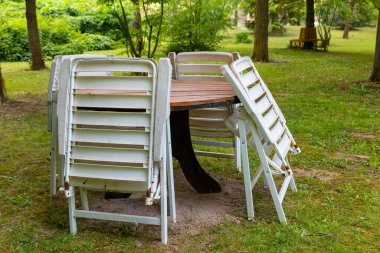 Huzurlu bir park ortamında ahşap bir masanın etrafında istiflenmiş sandalyeler. Yeşillik sakin bir atmosfer yaratıyor. Bu da açık havada yeni bir yemek deneyiminin tadını çıkardığını gösteriyor.
