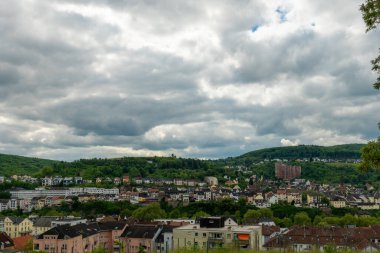 Fırıl fırıl dönen bulutlarla dolu bir gökyüzünün altında yuvarlanan yeşil tepelerin arasına yerleştirilmiş pitoresk bir kasaba ortaya çıkıyor. Hareketli binalar manzarayı dolduruyor, barışçıl bir geri çekilme yaratıyor..