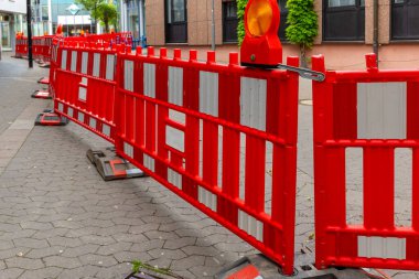 Parlak kırmızı güvenlik bariyerleri kaldırım taşı caddesinde devam eden onarım çalışmalarını gösteriyor. Şehir binaları arka planda yükselir, açık bir günde canlı bir şehir atmosferini yansıtır..