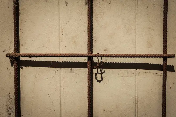 Yıpranmış metal çubuklar, güneşli bir duvarda karmaşık gölgeler oluşturarak, öğleden sonra sakin bir anda, engebeli doku ve yumuşak ışık arasındaki zıtlığı vurgular..