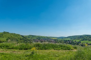Manzara boyunca uzanan canlı yeşil tepeler, açık mavi gökyüzünün altında yuva yapmış tuhaf bir köyü çerçeveliyor. Güneş yamaçlarda dans ederken doğa gelişir ve barışçıl bir atmosfer yaratır..