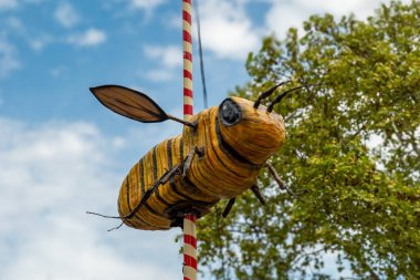 Açık hava festivalinde bir direkten sarkan canlı bir el işi arı süslemesi, yapraklarla çevrili ve parlak bir gökyüzünün altında kabarık bulutlarla sarkıyor. Neşeli atmosfer herkesi kutlamaya davet eder..