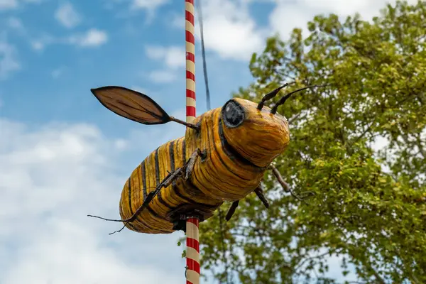 Açık hava festivalinde bir direkten sarkan canlı bir el işi arı süslemesi, yapraklarla çevrili ve parlak bir gökyüzünün altında kabarık bulutlarla sarkıyor. Neşeli atmosfer herkesi kutlamaya davet eder..