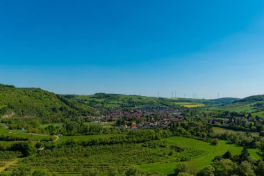 Canlı yeşil manzaraların arasına yerleşmiş büyüleyici bir köye bakan bu sakin yer, açık güneşli bir gökyüzünün altında doğanın güzelliğini gösteriyor, uzaktaki rüzgar türbinlerini vurguluyor..