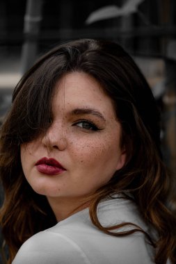 Sıcak güneş ışığı, genç bayanı nazikçe çarpıcı çillerle ve kalın dudaklarla sarar. Düşünceli bir şekilde uzaklığa bakarken şehir zemininin ortasında çekici bir atmosfer yaratır..