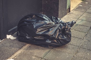 Sessiz bir şehir geçidinde, büyük siyah bir çöp torbası bir duvara yaslanır, öğleden sonra güneş ışığının yumuşak parıltısını yakalar. Yakında başıboş kağıtlar hafif meltemde sallanıyor..