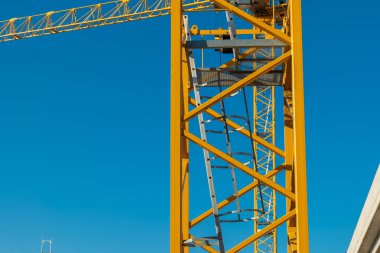 Parlak sarı bir vinç parlak mavi bir gökyüzüne doğru yükseliyor, inşa etmek için kullanılan karmaşık yapı ve tasarımı vurguluyor. Sahne, kentsel gelişimin özünü yakalıyor..