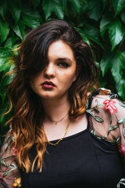 Genç bir kadın yoğun yeşil yaprakların önünde güven içinde duruyor. Koyu renk saçları omuzlarının üzerinden dökülüyor, elbisesindeki renkli nakışları vurguluyor..