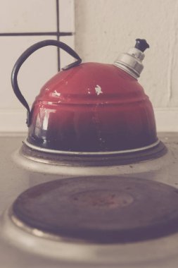 Eski moda kırmızı çaydanlık sobanın tepesinde oturur. Klasik cazibesi, yumuşak sabah ışığı altında parlar. Mutfak sessiz, su ısındıkça sıcak bir fincan çay için beklentiler artıyor..