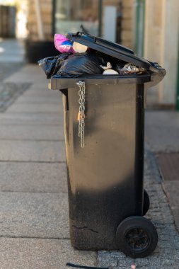 Dolu bir çöp tenekesi kalabalık bir kentsel alanda kaldırımda duruyor, güneş ışığı çöplerin üzerine yükseliyor. Yakındaki kaldırımda aşınma izleri görülüyor, kentsel yaşam ve zorluklar vurgulanıyor.