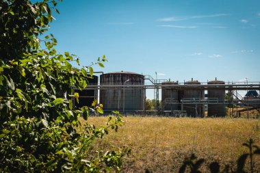 Uzun yeşil bitkiler, birkaç büyük endüstriyel tankın yer aldığı geniş bir araziyi çerçeveliyorlar. Parlak mavi gökyüzüne karşı hazırlanan bu kırsal alan, gündüz vakti doğa ve endüstrinin karışımını vurguluyor..