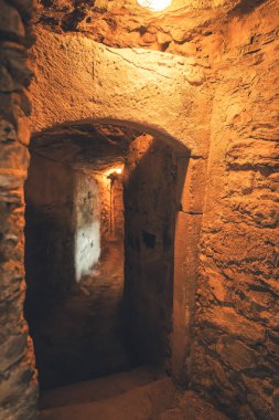 Işıl ışıl yanan bir geçit yıpranmış taş duvarlardan geçerek unutulmuş bir uzayın tarihini gözler önüne seriyor. Yumuşak ışık, kırsal, dünyevi dokuları vurgular, keşfi davet eder..