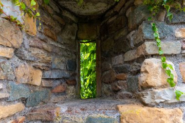 Güneş ışığı, yıpranmış taş duvarlardaki dar bir delikten süzülerek dışarıdaki yemyeşil yeşil alana hafif bir parıltı salıyor. Sarmaşıklar taşları sarar, bu gizli köşenin huzurlu atmosferini güçlendirir..