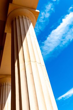 Uzun, dokulu sütunlar açık mavi gökyüzüne yükseliyor, karmaşık mimari tasarımı vurguluyor. Yapı tarafından dökülen gölgeler canlı sahneye derinlik ekler.