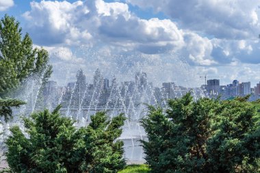 Şehir parkında geniş nehre karşı dans eden yaya çeşmesi. Zemin kemer bazlı fıskiyeler arka planda modern şehir manzarasında akar. Büyük evler ve binalar su sıçratarak ve su damlatarak görülebilir..