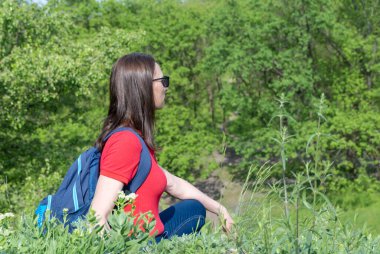 Sırt çantalı turist kadın gergin oturup yaprak döken ormanlara bakıyor. Yeşil Vadi 'de aktif bir yetişkin yürüyüşü. Gezgin şehvetin ve bahar çimenli tepenin tadını çıkar. Dişi yürüyüşçü keşfi.