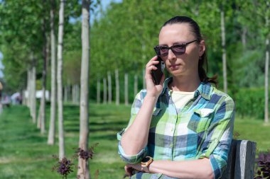 Parktaki bankta oturan ve cep telefonuyla konuşan bir kadın. Genç yetişkinler sohbet etmek için akıllı telefon kullanır. Kadın dinleniyor ve cep telefonuyla konuşuyor. Boş zaman, teknoloji ve modern yaşam tarzı kavramı.