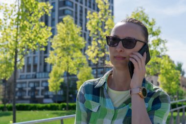 Park yerinde dinlenen ve cep telefonuyla konuşan bir kadın. Genç yetişkinler sohbet etmek için akıllı telefon kullanır. Kadın dinleniyor ve cep telefonuyla konuşuyor. Boş zaman, teknoloji ve modern yaşam tarzı kavramı.