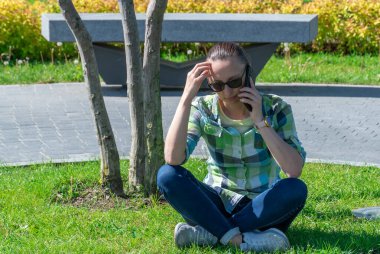 Park çimlerinde oturan ve cep telefonuyla konuşan bir kadın. Genç yetişkinler sohbet etmek için akıllı telefon kullanır. Kadın dinleniyor ve cep telefonuyla konuşuyor. Boş zaman, teknoloji ve modern yaşam tarzı kavramı.