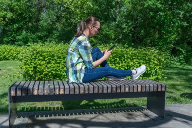 Parktaki bankta oturan ve cep telefonuyla ekranı çeviren kadın. Genç yetişkinler sohbet etmek için akıllı telefon kullanır. Parmaklar telefon ekranına dokunur. Kadın dinlenme ve cep telefonuyla iletişim.