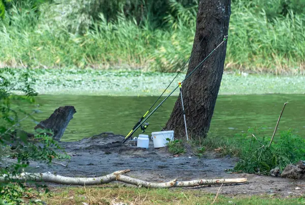 stock image Fishing tackle and fish container on river bank. Ready to go fishing sunrise in secluded place. Fishing pole and spinning rod getting away from it all. Rural getaway and active hobby in summer day.