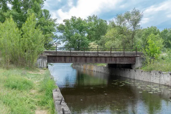 stock image Iron concrete bridge over the river canal. Reinforced pedestrian from one bank of water to other. Picturesque trees and bushes grow along the support slabs. Empty footbridge of bridge-canal with pier.