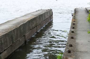 Pahalı dar nehir kanalı boyunca destek levhaları. Demir beton iskeleyi bir su bankasından diğerine güçlendirdi. Ağaçlar ve sazlıklar boş yayalar boyunca büyürler. Nehirdeki yansıma doğası.